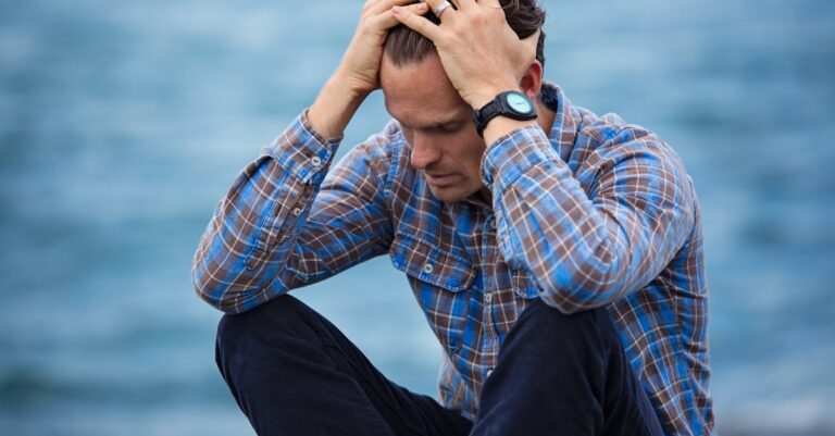 A man in a plaid shirt sits by the water looking distressed, symbolizing stress.